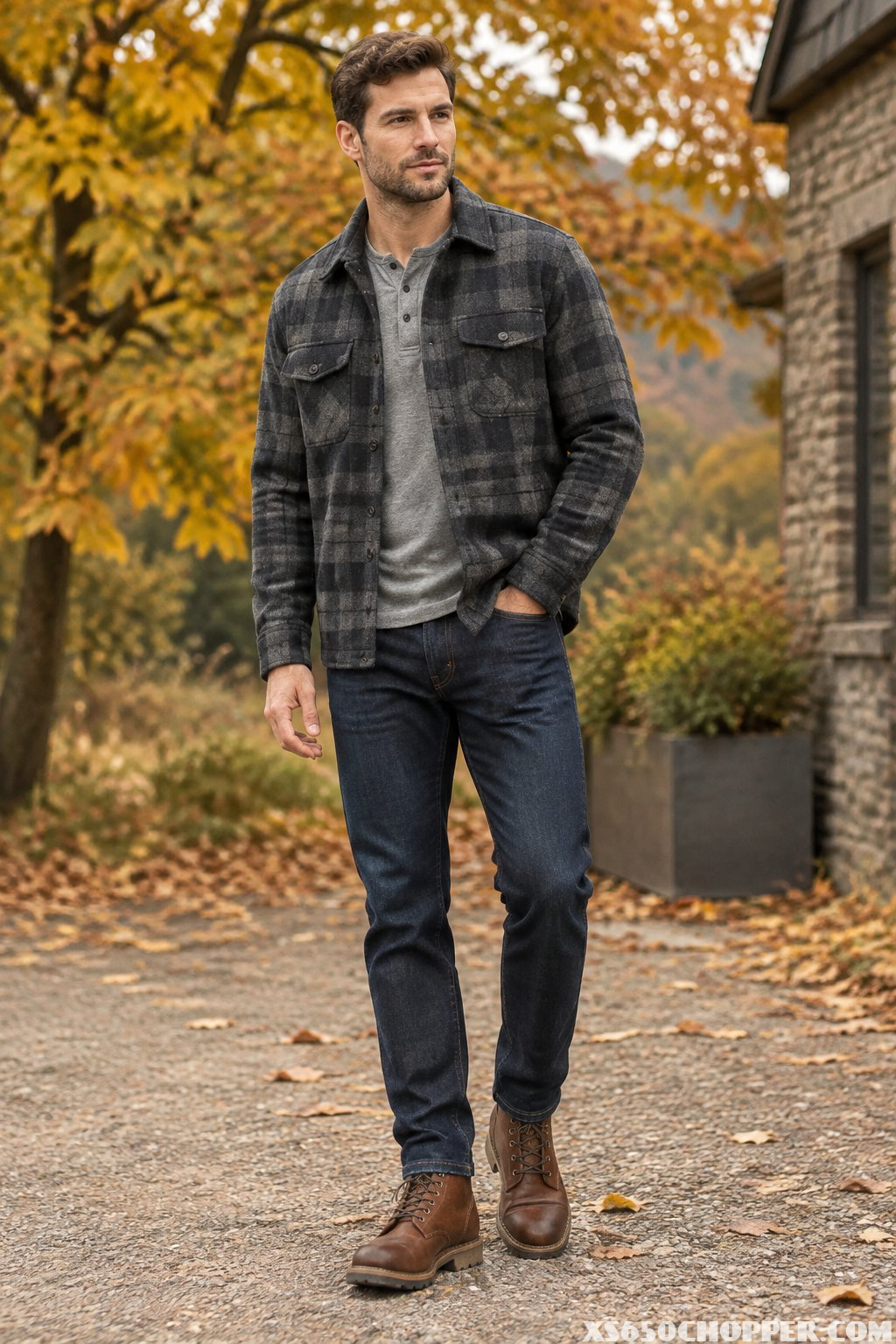A man wearing a plaid wool overshirt, gray henley, indigo jeans, and brown work boots outdoors in autumn.