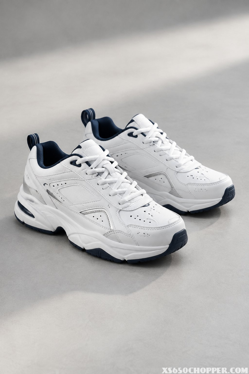 A pair of chunky white sneakers with silver and navy accents on a light gray floor.