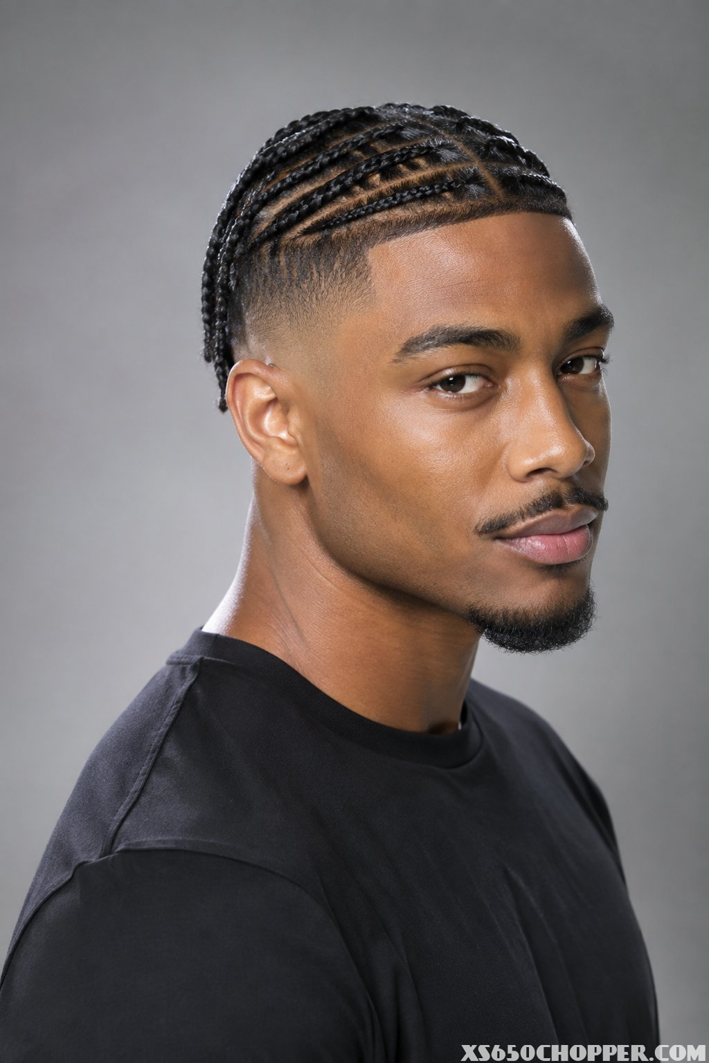 A man with small braids and a faded haircut poses against a plain background.
