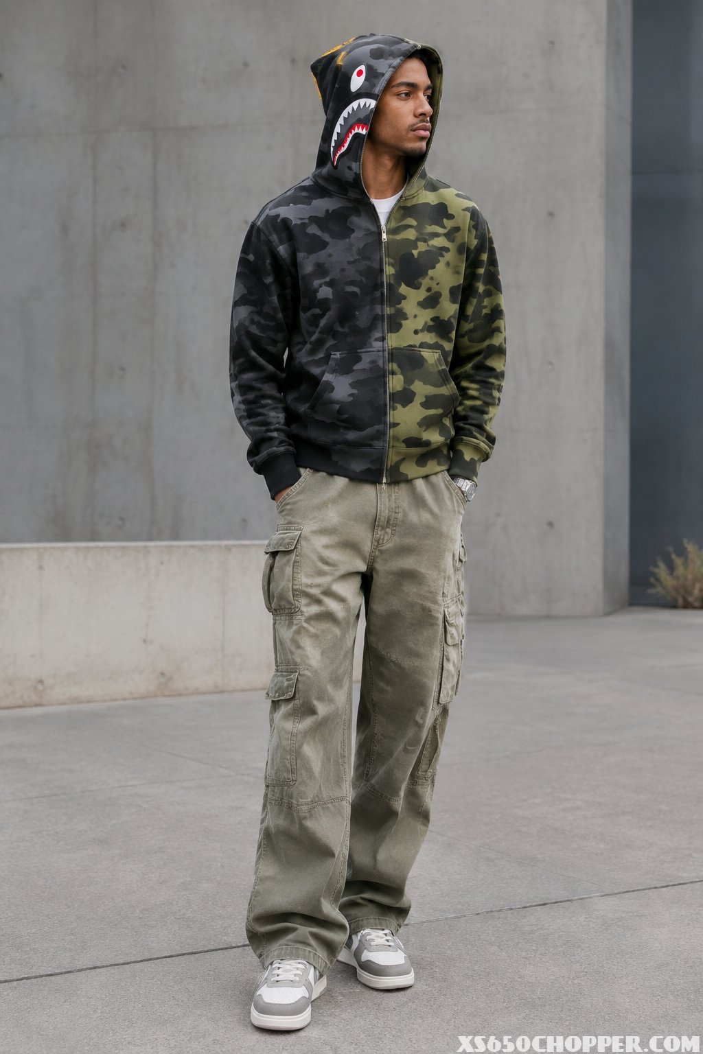 A man stands outdoors wearing a shark hoodie, baggy carpenter pants, and sneakers.