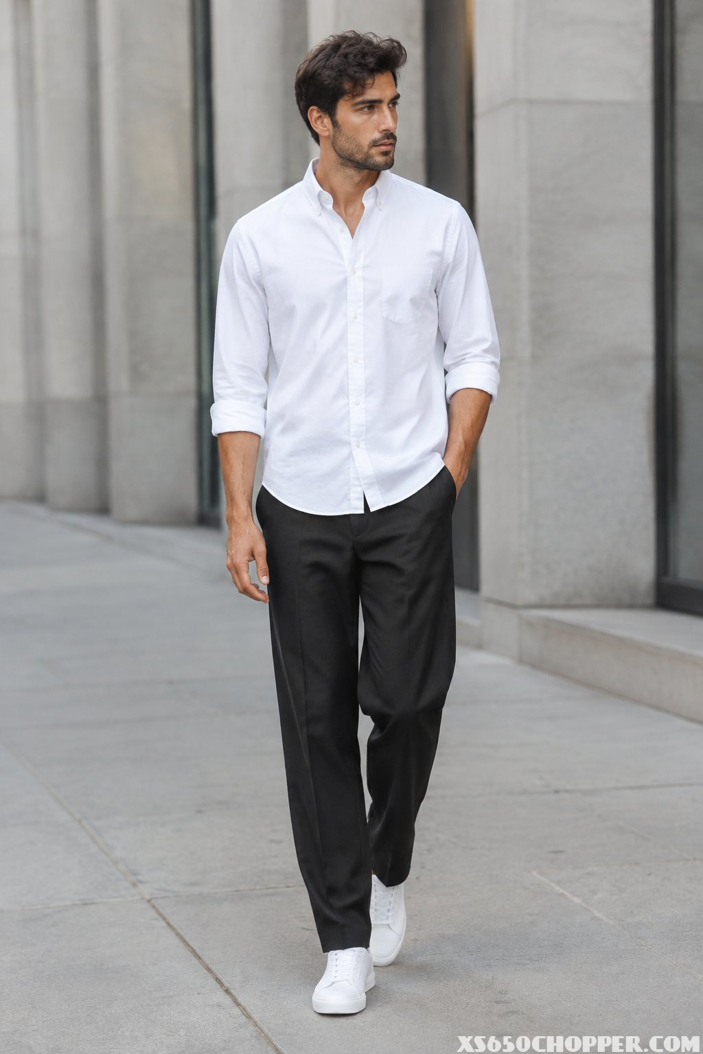 A man wearing a white button-down shirt, relaxed black trousers, and white sneakers stands on a city sidewalk.