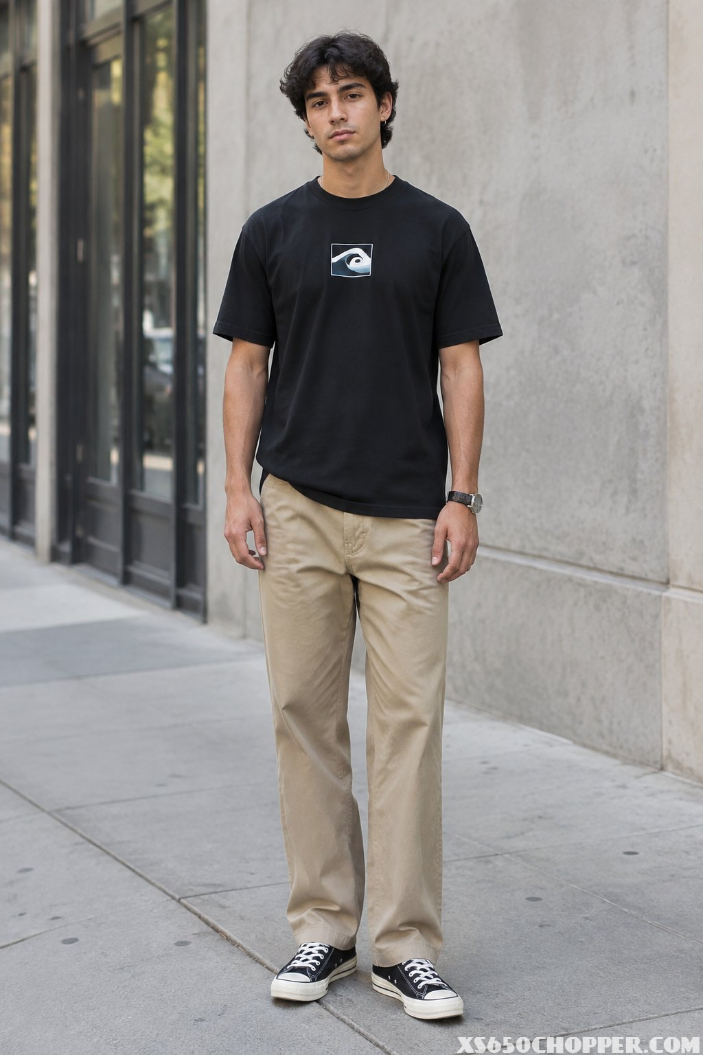 A man stands on a city sidewalk wearing a black T-shirt, khaki work pants, and black-and-white sneakers.