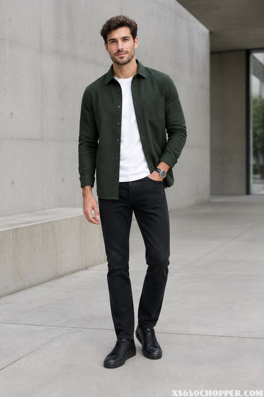A man wearing a dark green overshirt, white T-shirt, and black jeans stands in an urban setting.