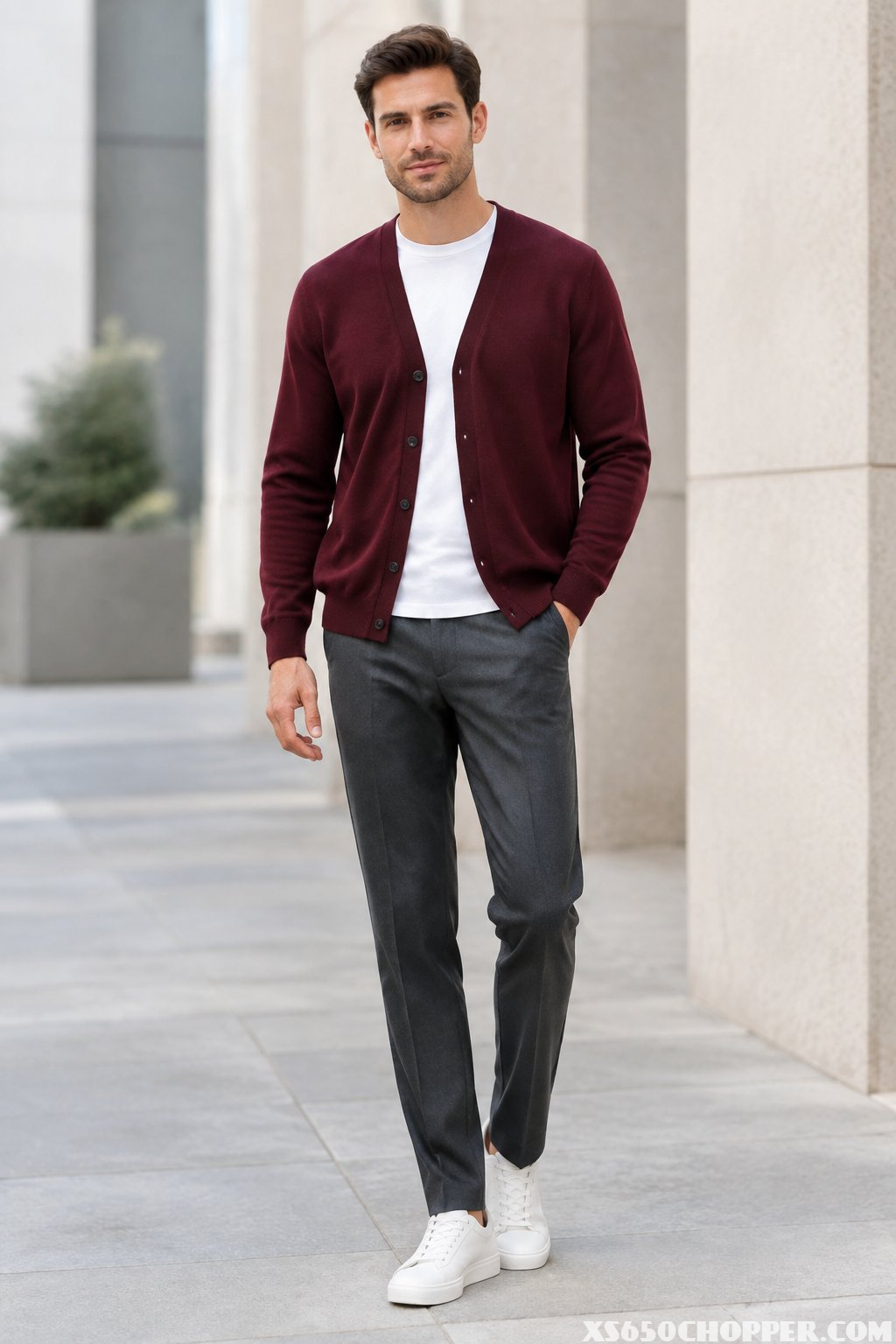 A man wearing a burgundy cardigan, white T-shirt, and tailored gray trousers stands in a modern urban setting.