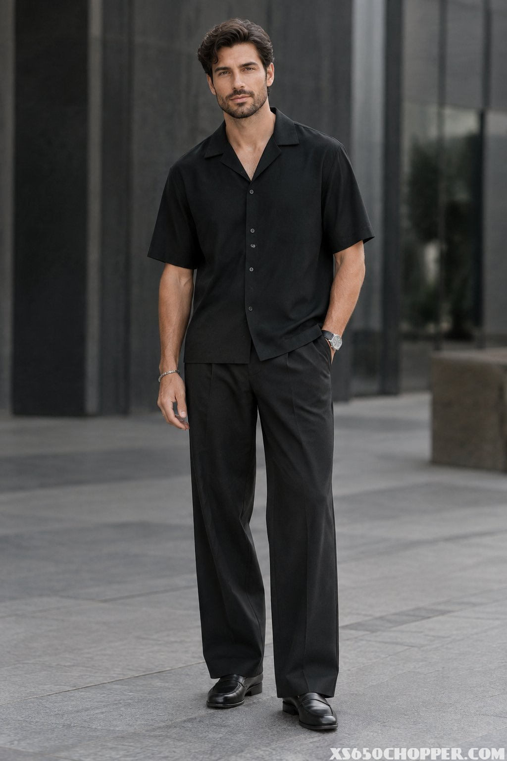 A man wearing a black camp-collar shirt and wide-leg trousers stands in a modern urban setting.