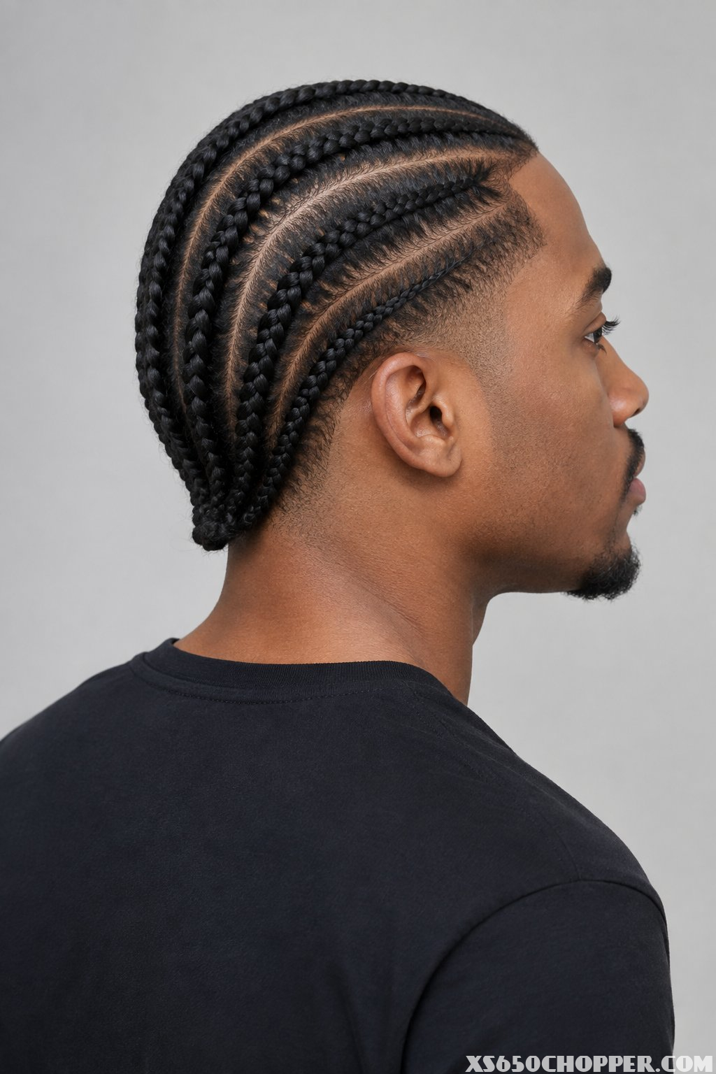 A man with straight-back cornrows and a low taper fade poses against a plain background.