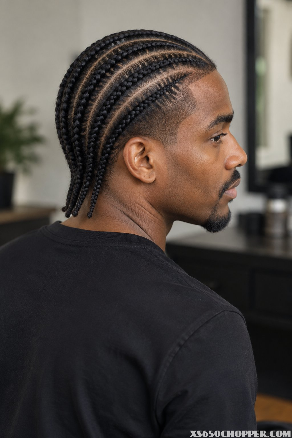 A man with neat straight-back braids sits against a softly blurred neutral background.