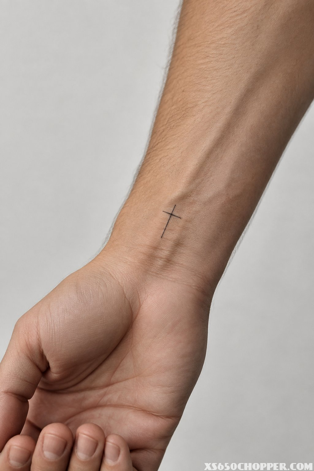 A close-up of a forearm showing a small fine-line cross tattoo on the inner wrist.