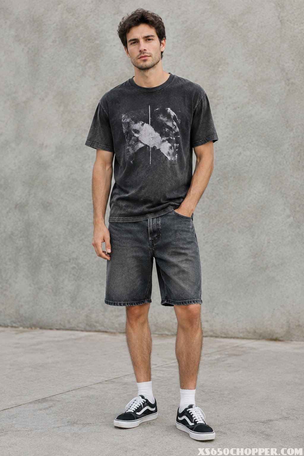 A man wears a washed black graphic T-shirt, charcoal denim shorts, and black-and-white sneakers in an urban setting.