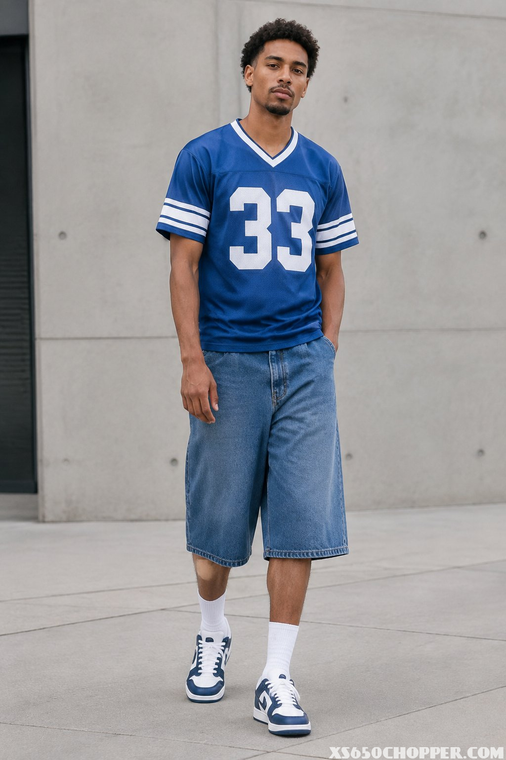 A man wearing a vintage sports jersey, long denim shorts, white socks, and low-top sneakers stands on a city street.
