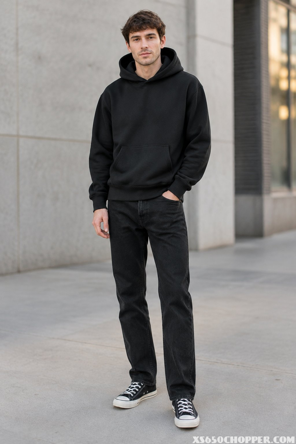 A man wearing a black hoodie, black jeans, and black-and-white sneakers stands in an urban setting.