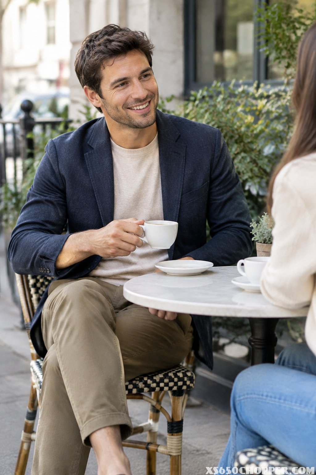 A man in a blazer, knit shirt, and chinos shares coffee at an outdoor café.