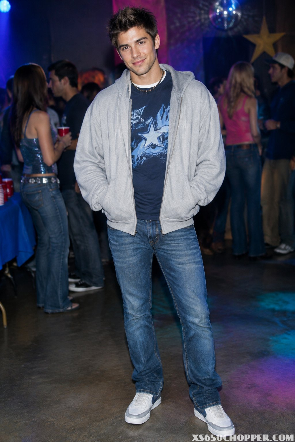 A man wears an open zip-up hoodie over a graphic T-shirt with skinny jeans at a party.