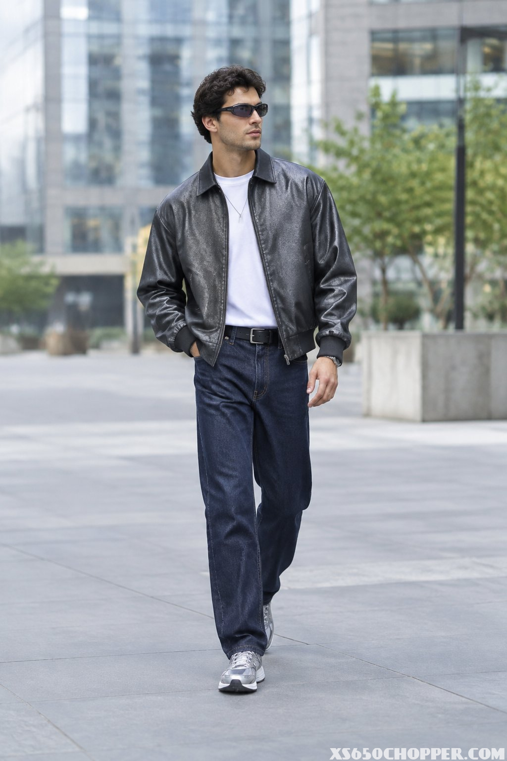 A man walks along a city street wearing a jacket, jeans, and sneakers.