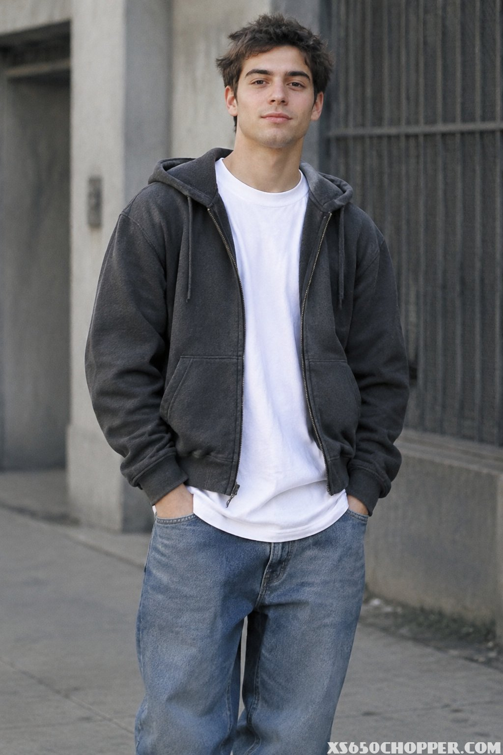 A man stands outdoors wearing a zip hoodie over a long T-shirt and jeans.