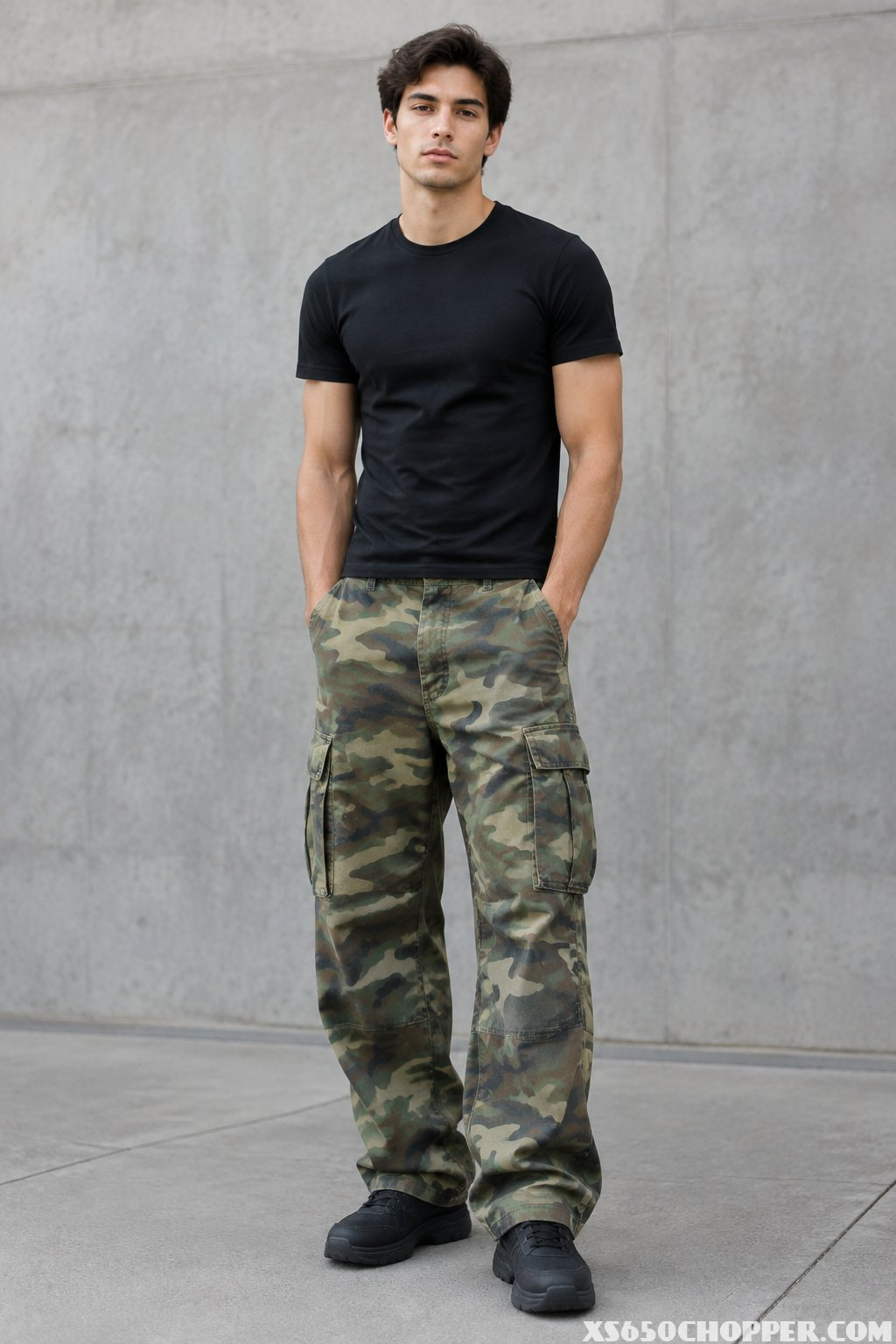 A man stands against a concrete wall wearing a black T-shirt and camouflage cargo pants.