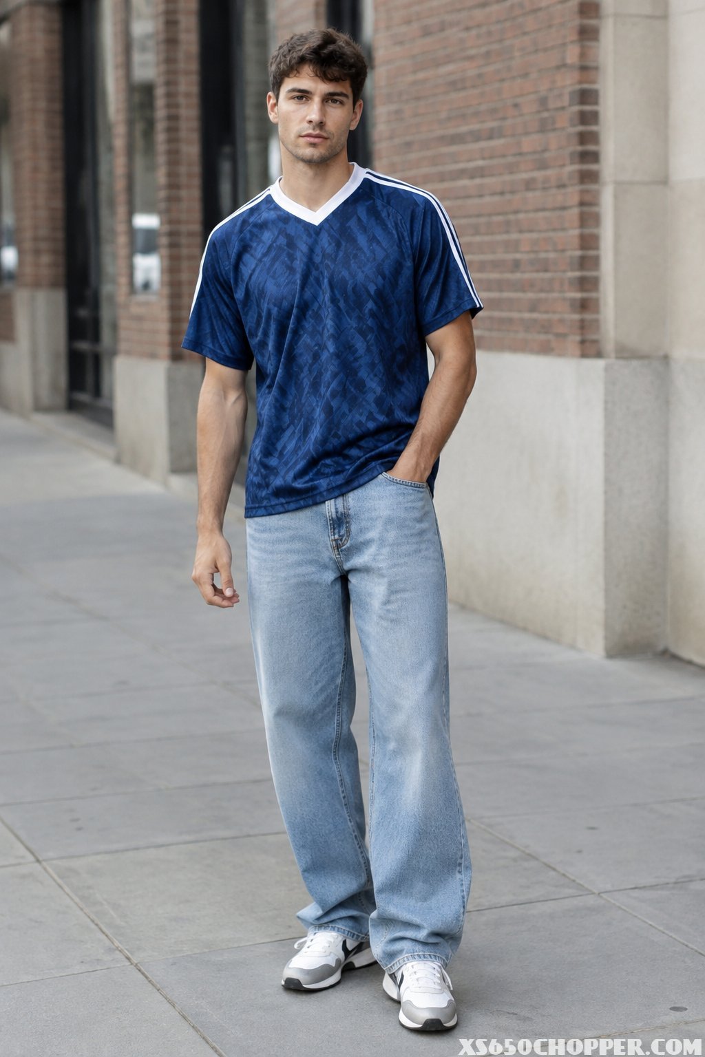 A man stands on a city sidewalk wearing a soccer jersey, baggy jeans, and trainers.
