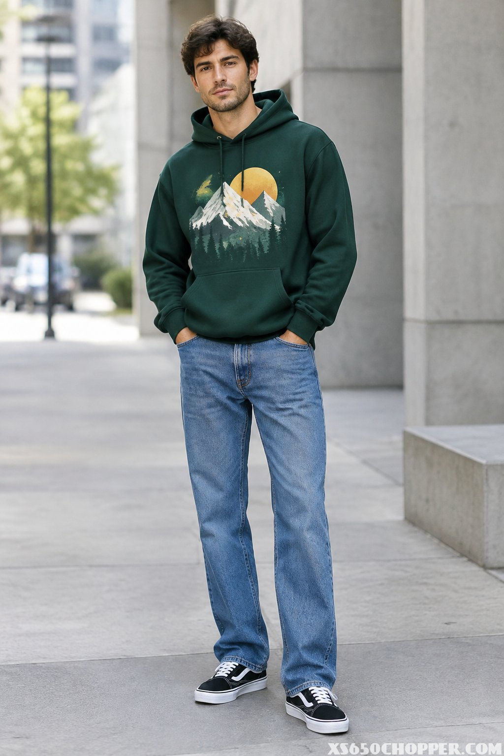 A man stands on a city sidewalk wearing a hoodie, jeans, and skate shoes.