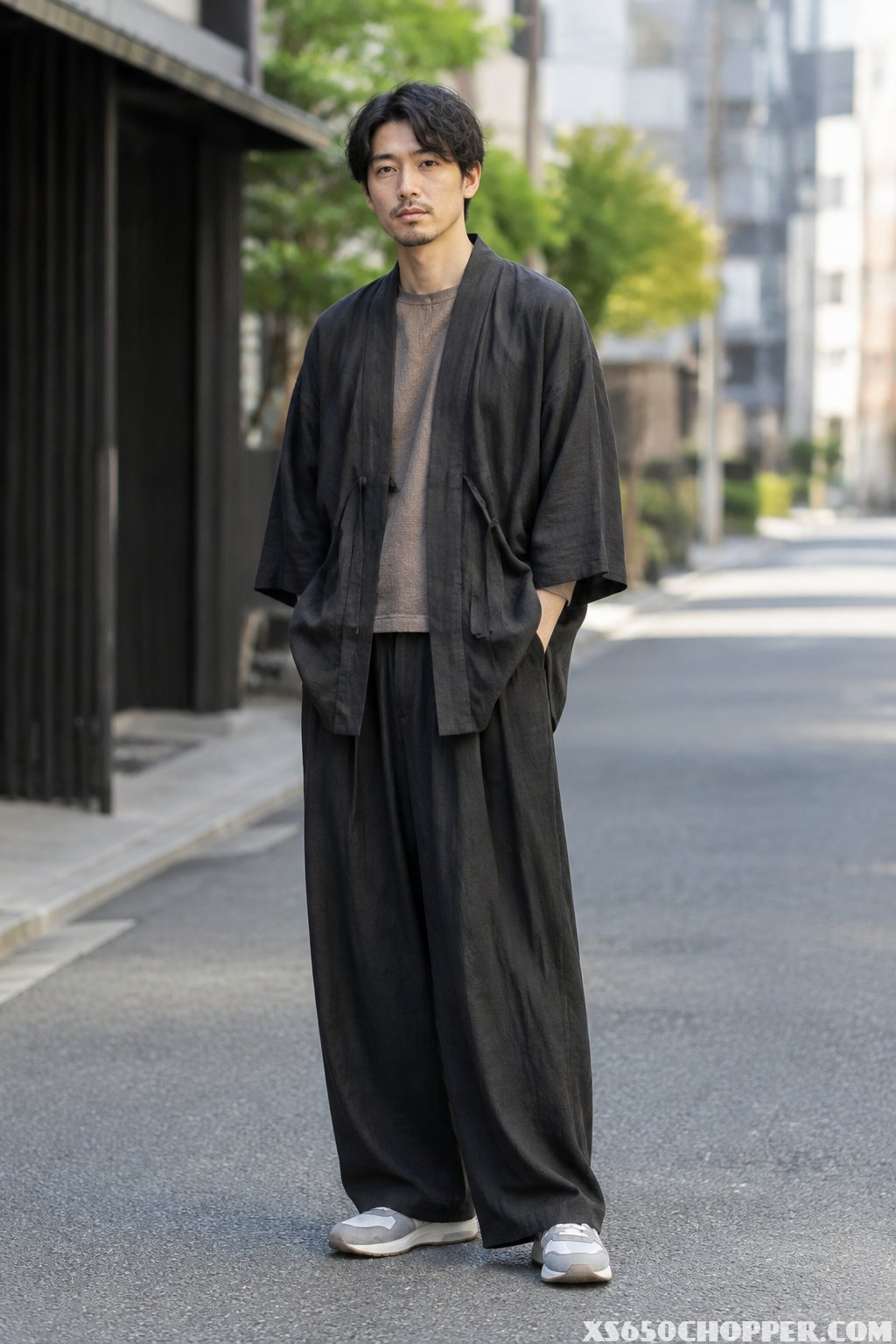 A man stands on a city street wearing a jacket and wide trousers.