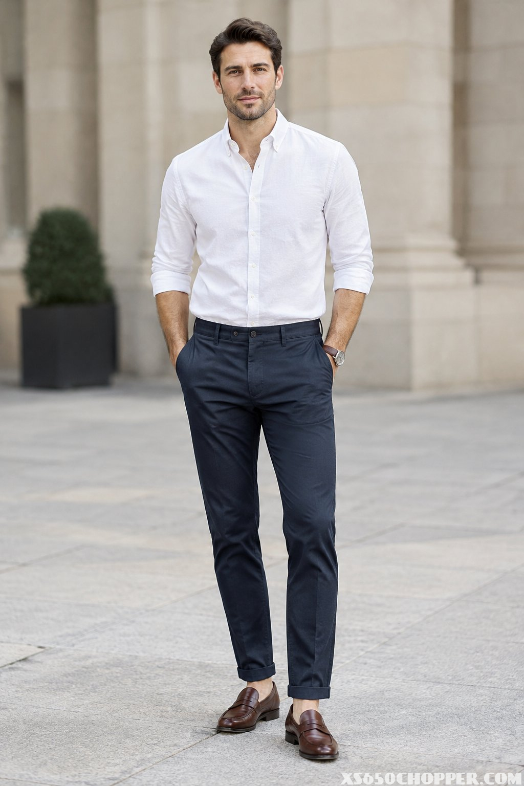 A man stands wearing a white shirt, navy trousers, and brown loafers.