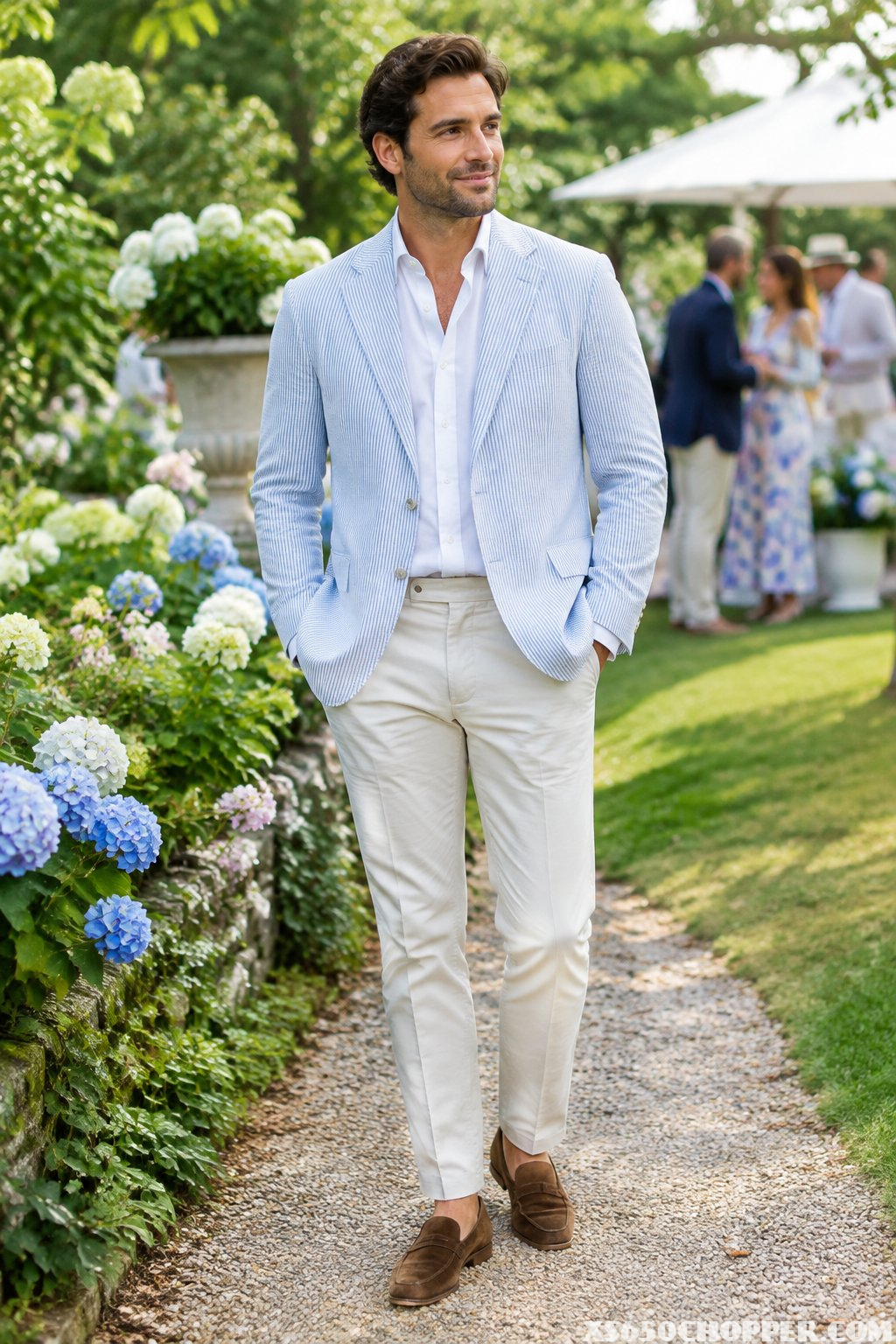 A man in a light jacket and chinos stands at a garden party.