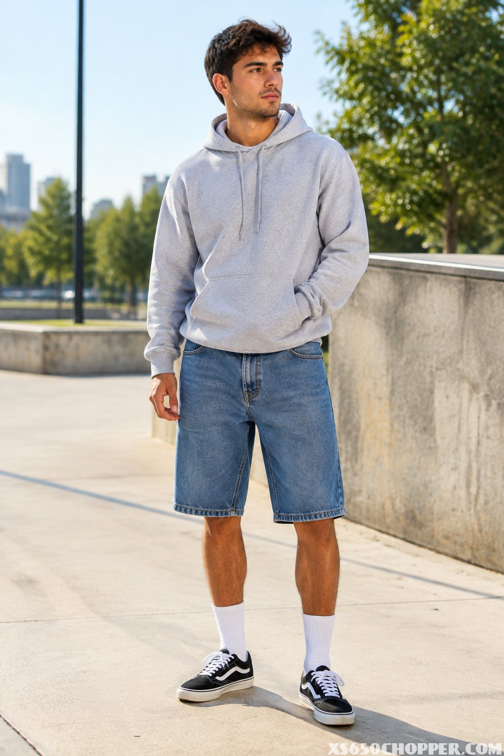 A man wearing denim shorts, a hoodie, crew socks, and skate shoes stands on a sidewalk.